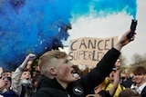 A young male in a crowd holds up a flare eminating blue smoke. A sign behind him reads "CANCEL SUPERLEAGUE"