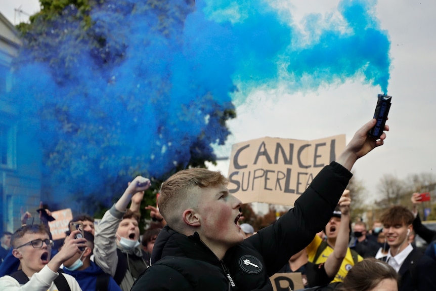 A young male in a crowd holds up a flare eminating blue smoke. A sign behind him reads "CANCEL SUPERLEAGUE"