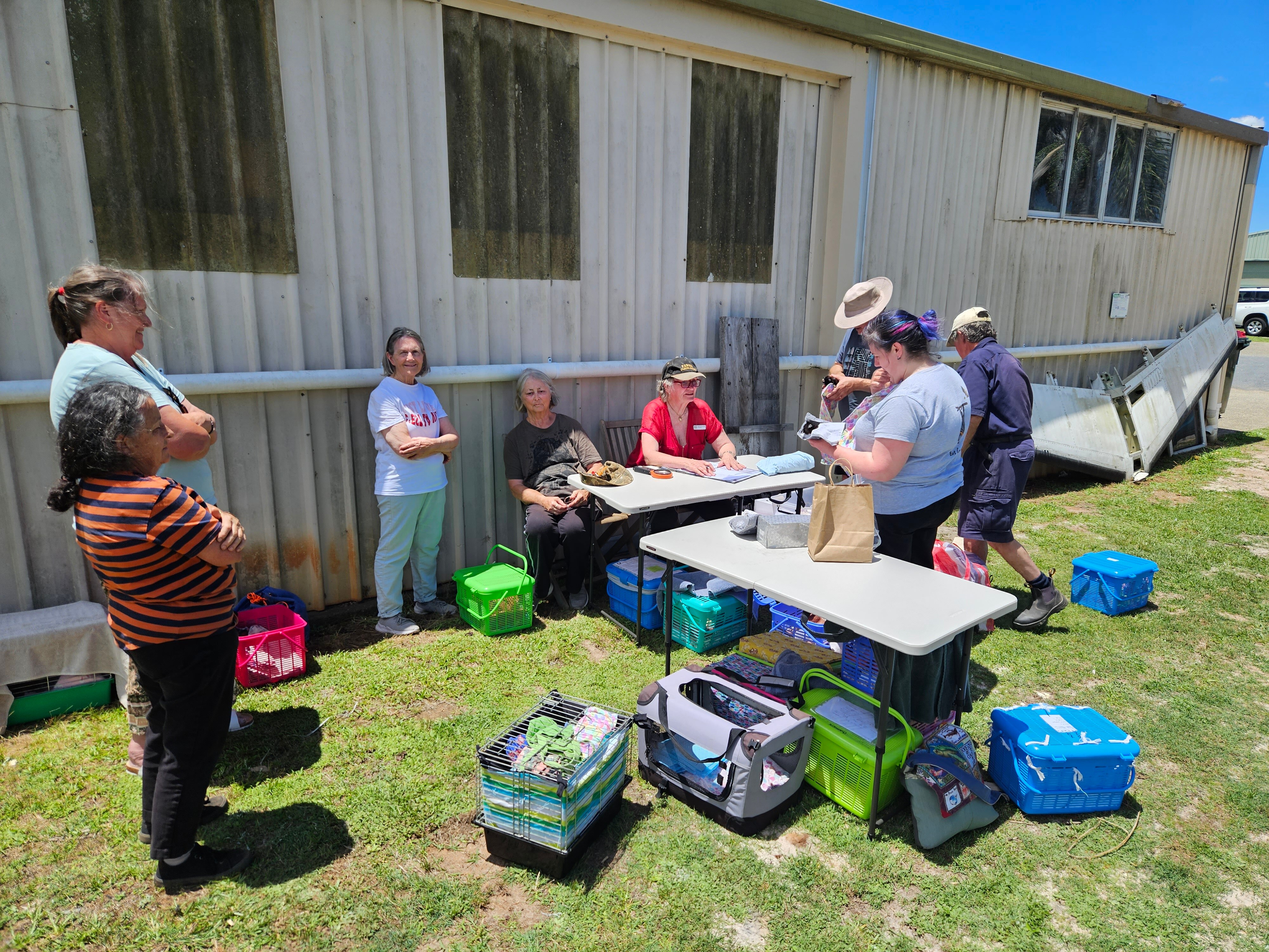 A group of people are standing nearby a shed. Some are sitting at foldout tables, others are standing.