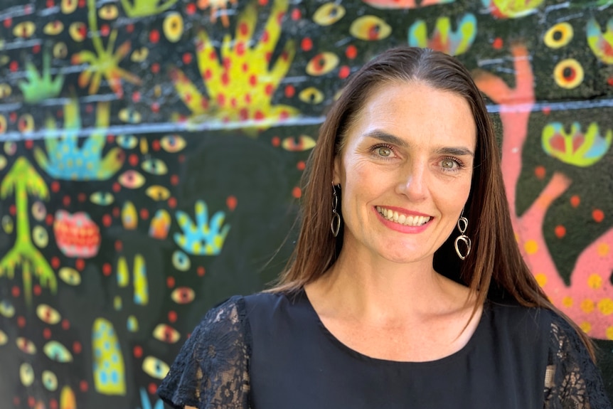 A woman with long hair smiles in from of a colourful mural