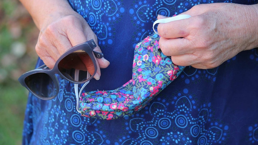 Close up of a woman's hands holding her sunglasses and her colourful homemade face mask.