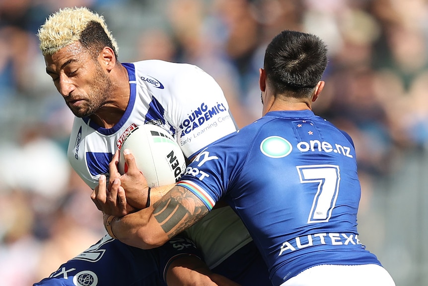 A Canterbury NRL player holds the ball as he is tackled by two Warriors opponents.