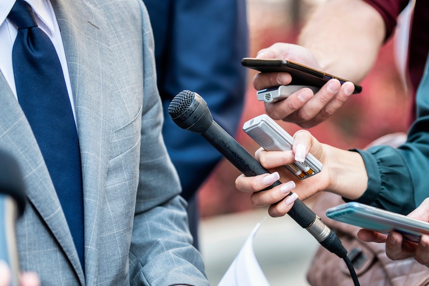 Journalists hold microphones in front of somebody speaking.