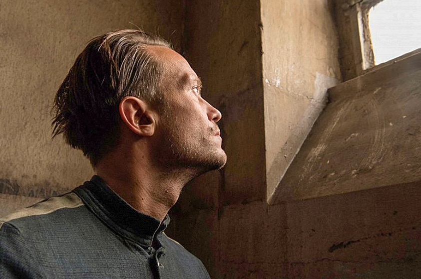 A man with short slicked back hair in prisoner attire looks up and is illuminated by daylight from small window is prison cell.