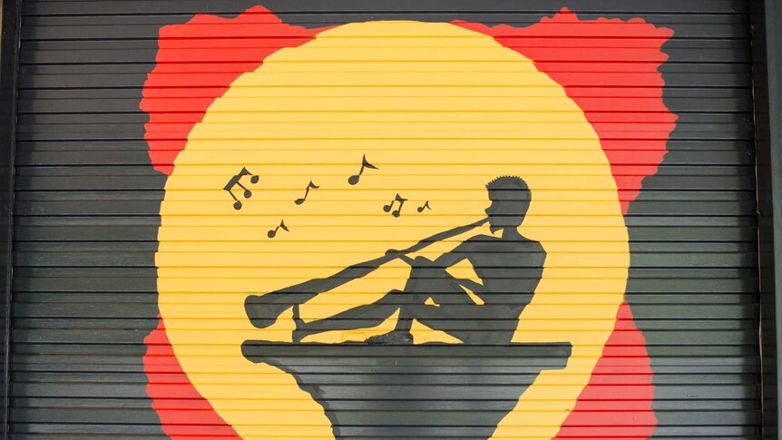 A silhouette painting incorporating the Aboriginal flag with a man playing a didgeridoo in the yellow section.