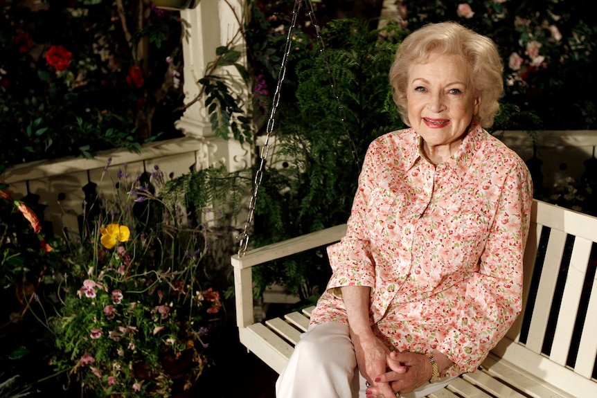 Betty white sitting on a white chair on a verandah wearing a pink blouse and white slacks