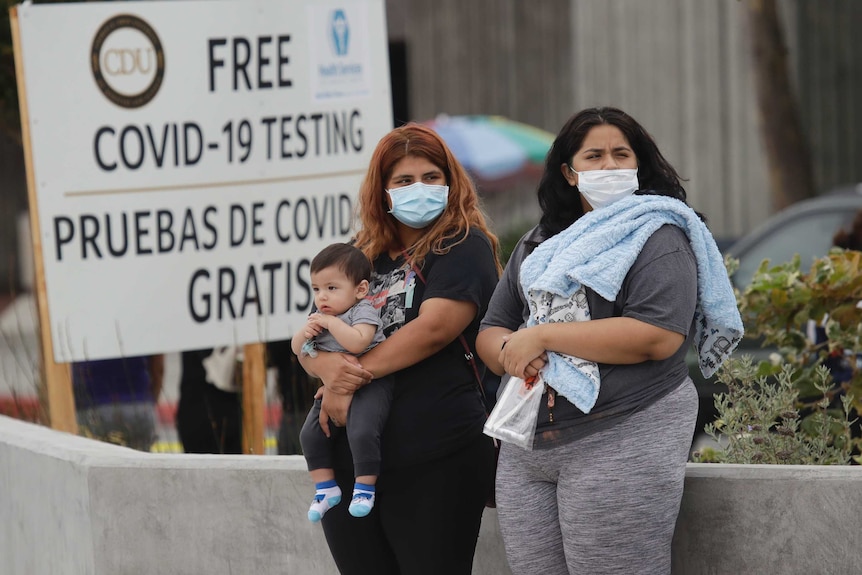 Two overweight Latin women in face masks, with one of them holding a baby, stand in front of a sign.