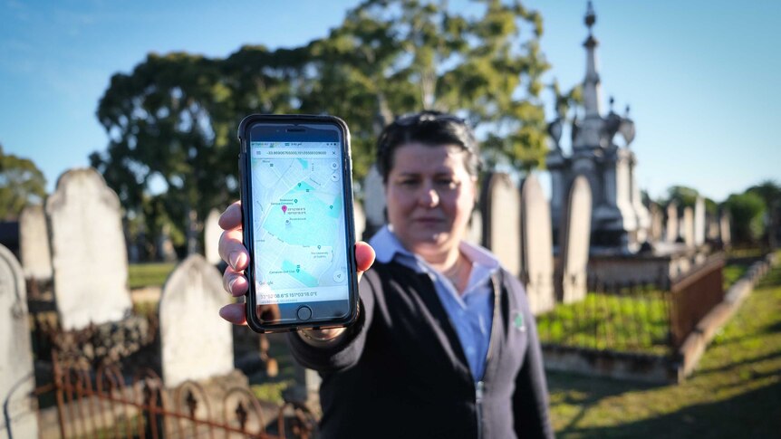 A woman holding Google Maps up, directing herself to a grave stone in Rookwood Cemetery.