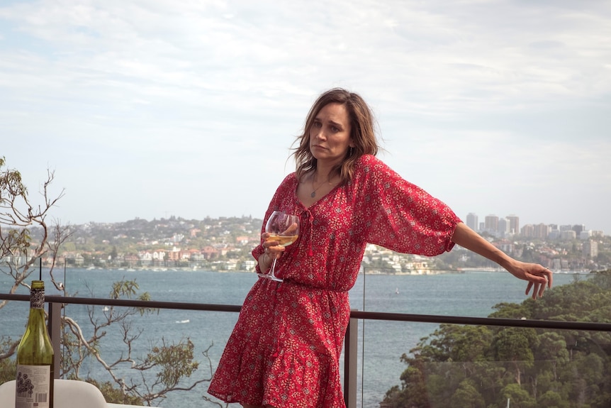Nicole da Silva as Simone looking sad with a wine in her hand, wearing a red dress standing on balcony against an ocean backdrop