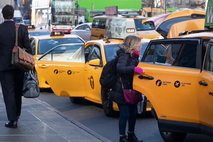 A woman in winter clothes holds open the door of a taxi cab, among a bunch of other taxis in New York City