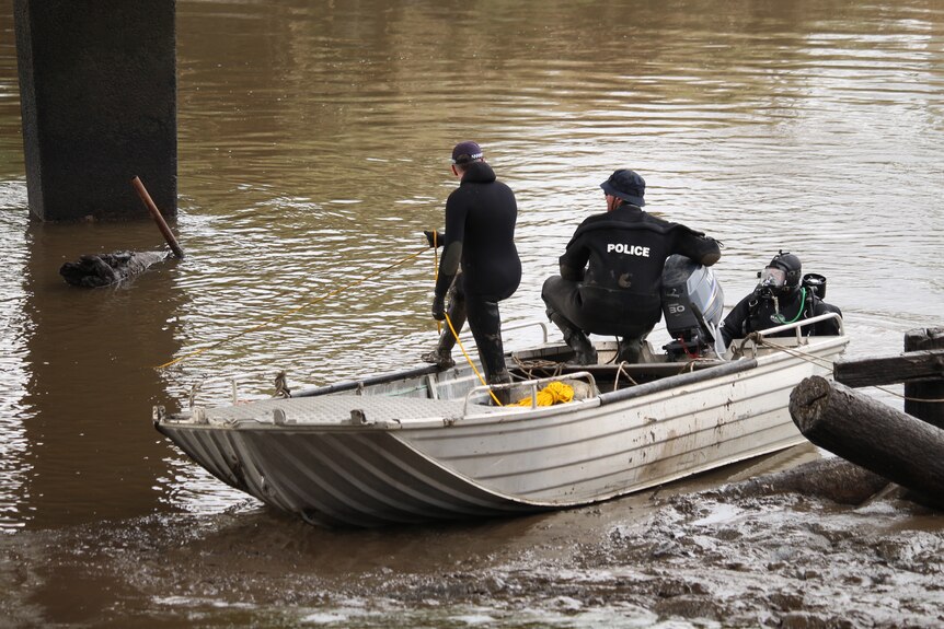 Two men in black wetsuits that say "police" on the back stand in a tin boat and look into a muddy river beneath a bridge.