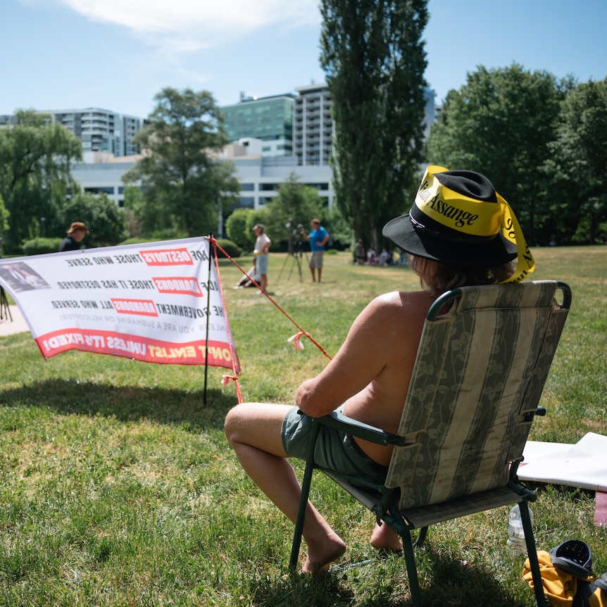 A shirtless man wearing a hat with a Julian Assange ribbon around it sits on a folding chair in a park. In the park is a stage.