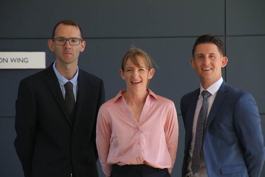 Three WA Police recruits stand smiling for a photo wearing plain clothes, a man on either side of a woman.