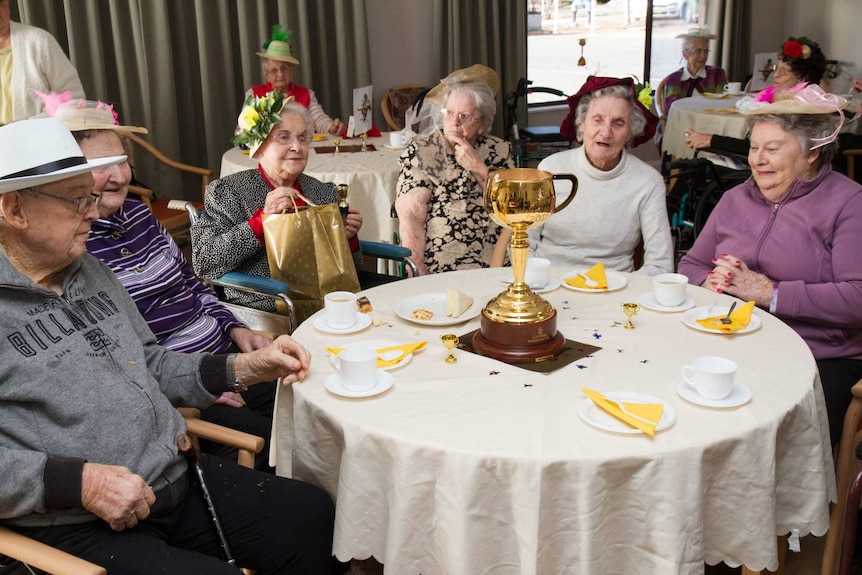 Older women and men sitting around a table wearing hats and looking at the gold Melbourne Cup trophy