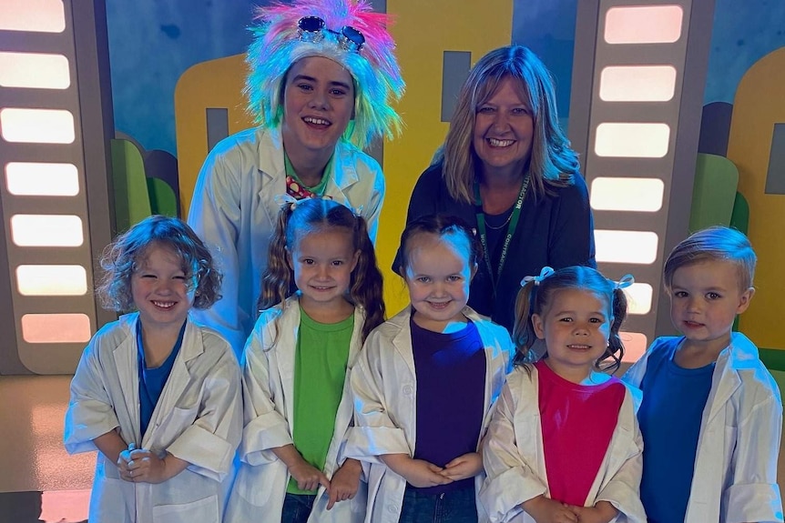 Two adults and five children in bright colours and white coats posing on a brightly coloured TV set.