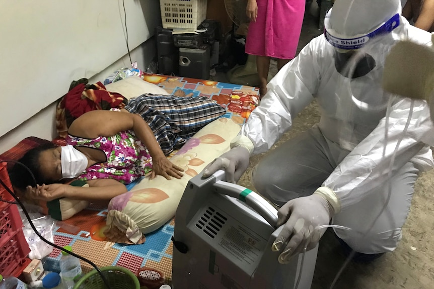 A man in full PPE crouches down next to a woman lying on a mat in a face mask