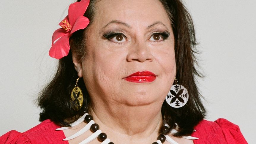 Head and shoulders image of an older Samoan woman in a bright red dress with islander jewelry and a hibiscus in her hair.