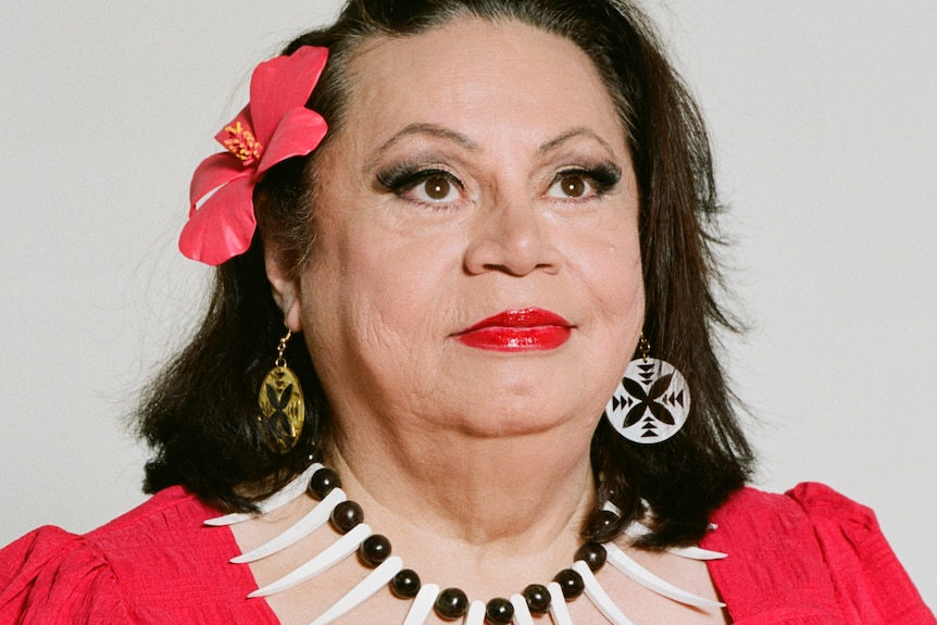 Head and shoulders image of an older Samoan woman in a bright red dress with islander jewelry and a hibiscus in her hair.