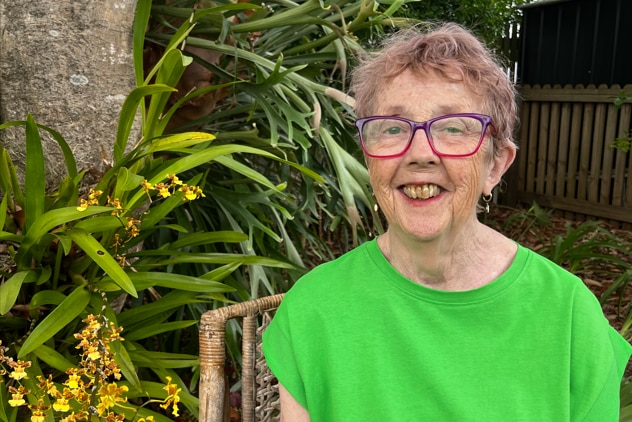 a lady wearing a bright green shirt sitting in a wooden chair in her garden smiling at the camera
