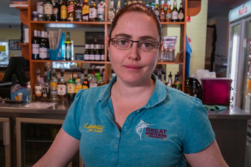 A woman stands in front of a bar full of spirits.