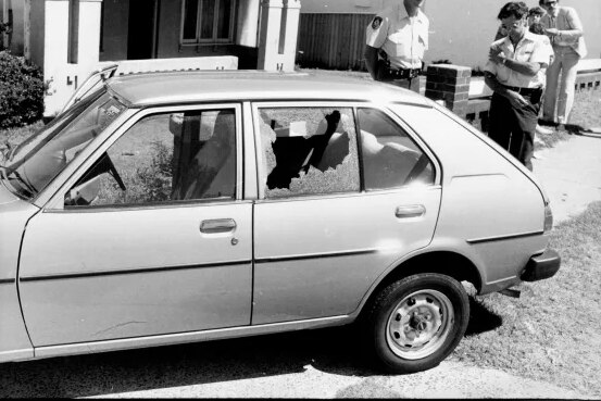 A black and white image of a car with boht windows shattered as police standby 