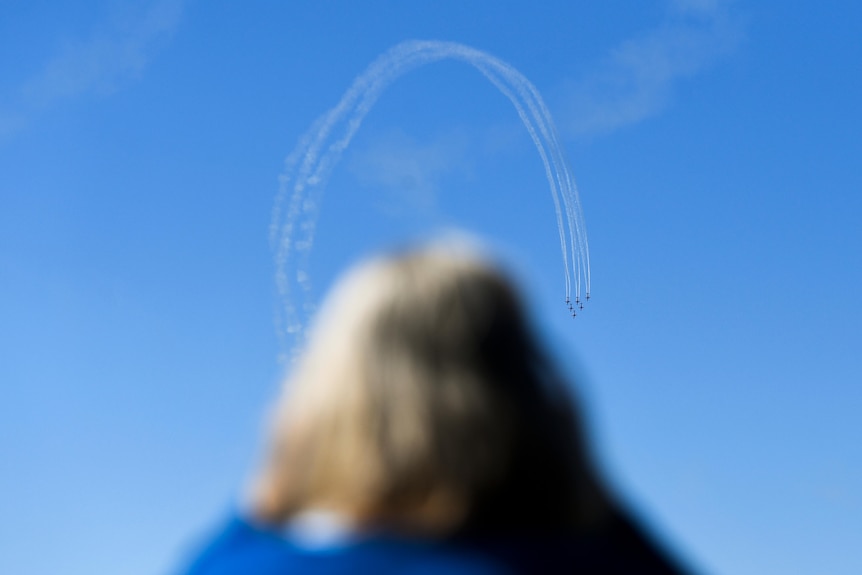 A woman watches planes fly in a loop, the smoke forming a curved trail through the air.