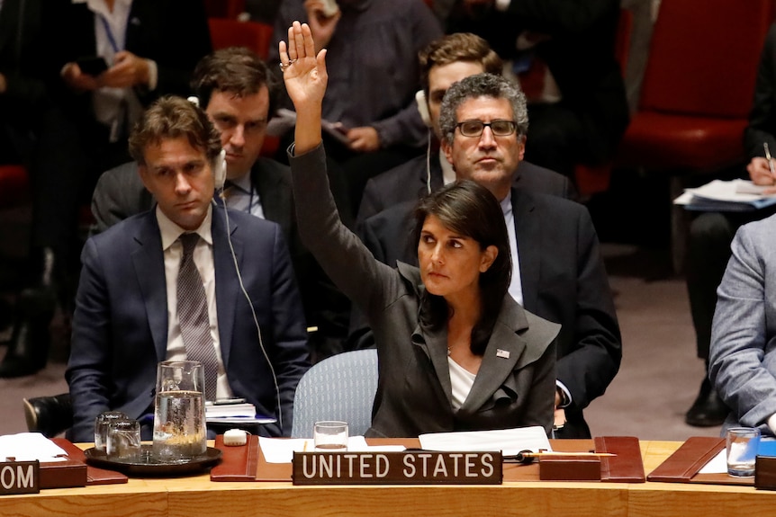 A woman in a grey suit sits with her hand raised behind a sign that says "United States"