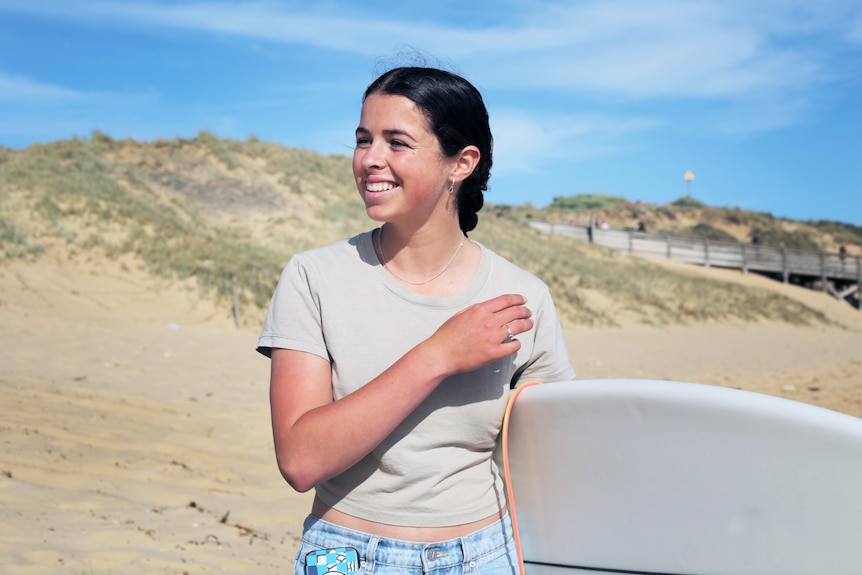 A teenage girl smiling, with a surfboard tucked under her arm.