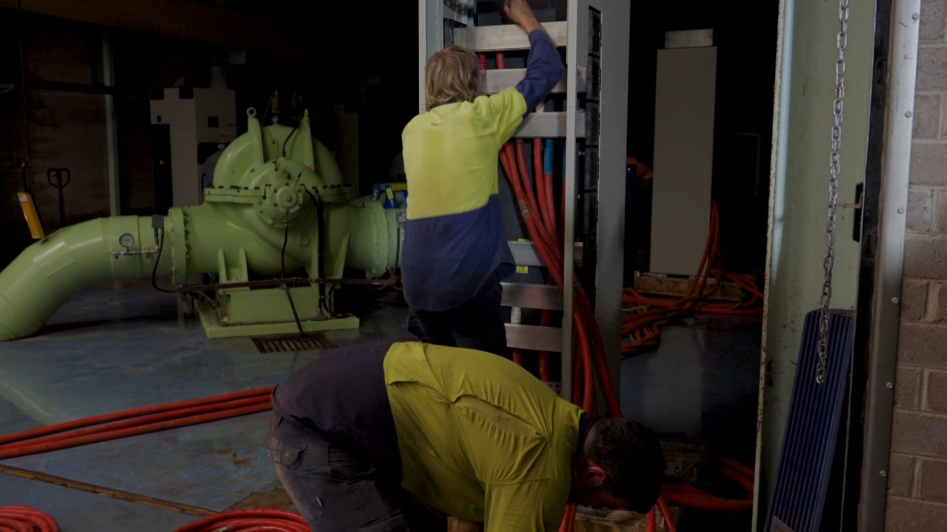 Two men in high-vis yellow uniform working in an industrial indoor space. One is leaning over metal shelving, red cords around