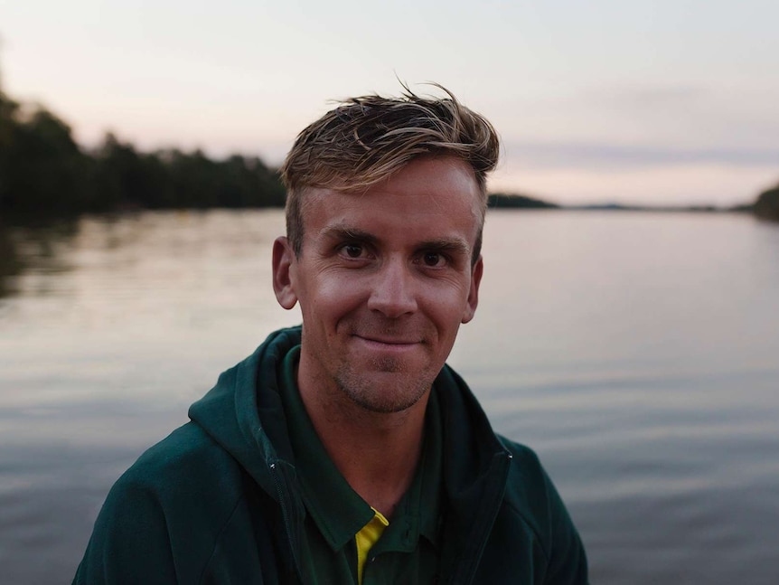 A man wearing a green jumper looks straight at the camera, river and trees in background at sunset.