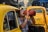 A taxi driver raises a bottle and pours a stream of water into his mouth as he sits in his car and leans outside.