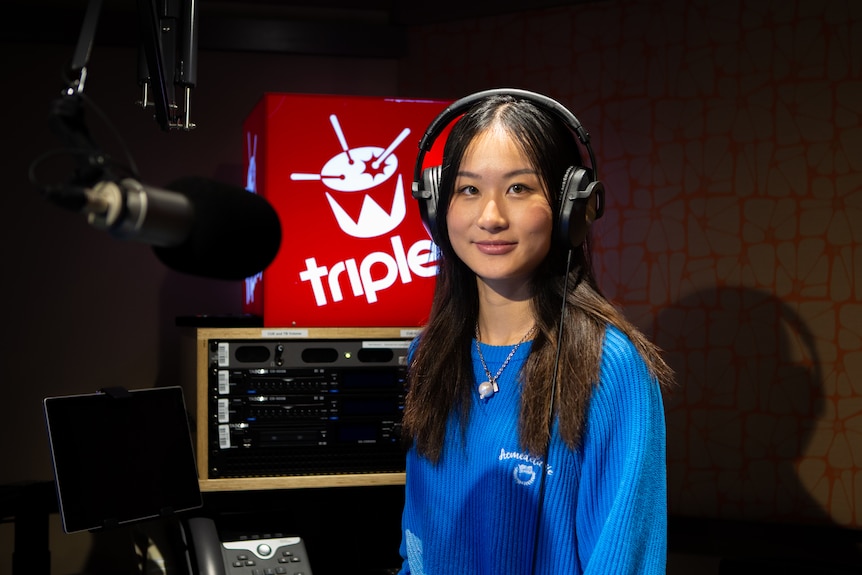 Girl wears blue jumper and headphones and looks to the camera smiling. A red box with triple j and a drum in white is lit up.