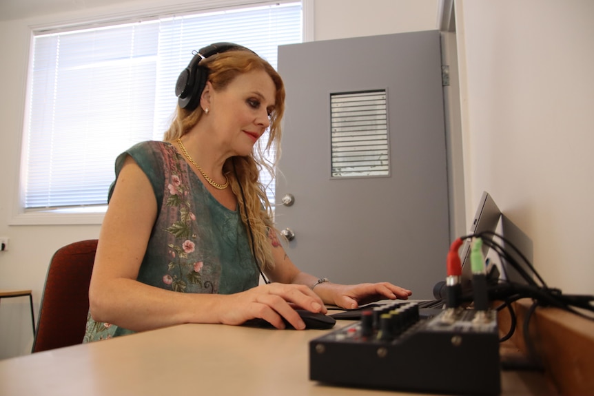 Una mujer con el pelo largo y rojo y una blusa colorida con audífonos sentada en un escritorio con una computadora portátil y un equipo de sonido