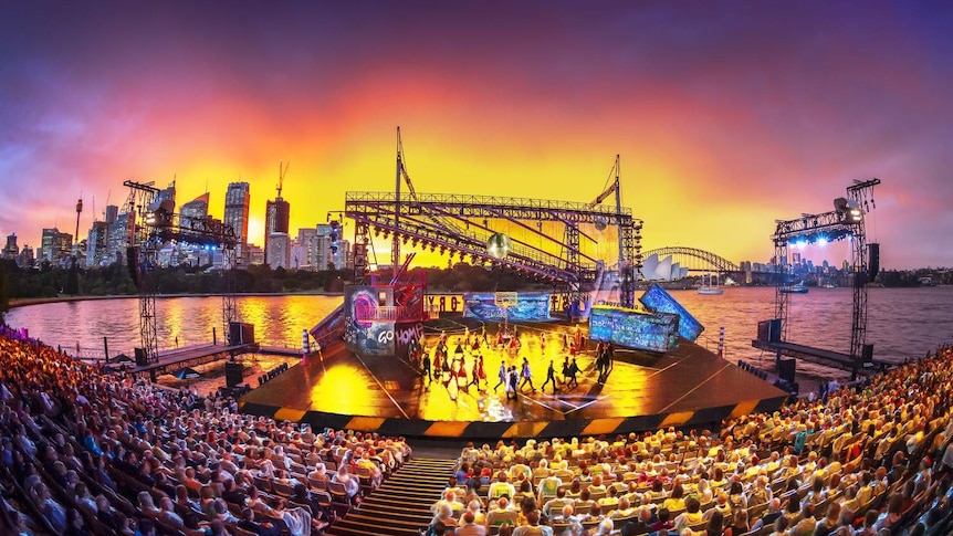 An audience seated in front of actors on stage, with a brightly coloured sunset over Sydney Harbour in the background.