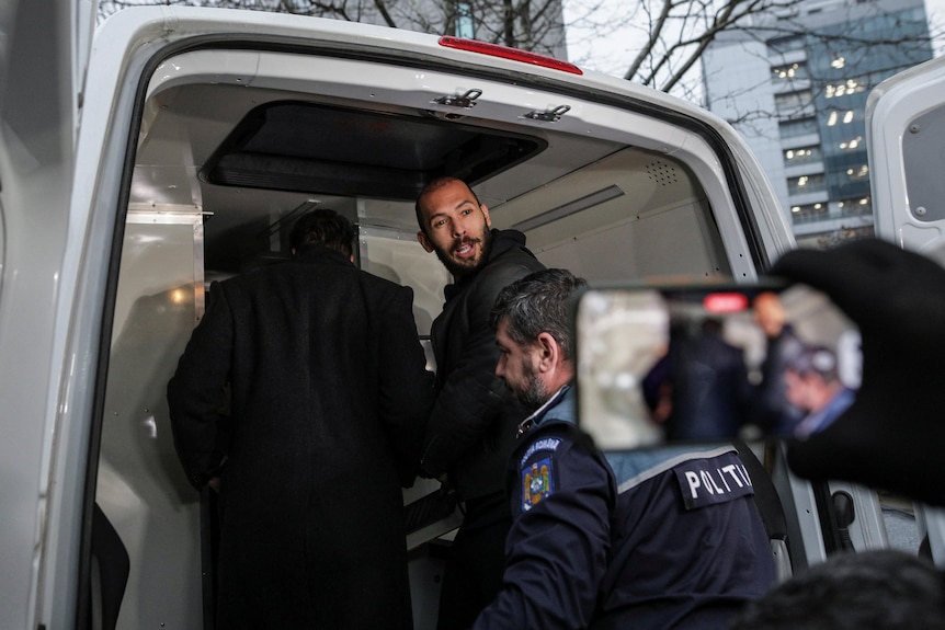 A police officer helps two men into the back of a van as someone captures the image on a mobile phone