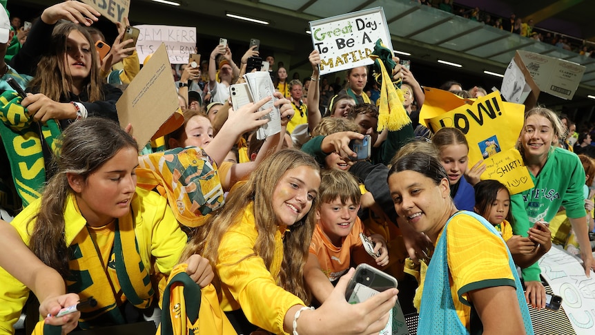 A woman soccer player wearing a yellow and green jersey with a blue bib poses for a photo with fans wearing yellow in a crowd