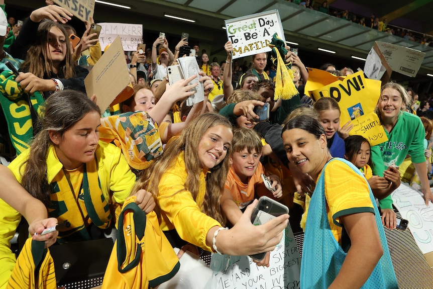 A woman soccer player wearing a yellow and green jersey with a blue bib poses for a photo with fans wearing yellow in a crowd