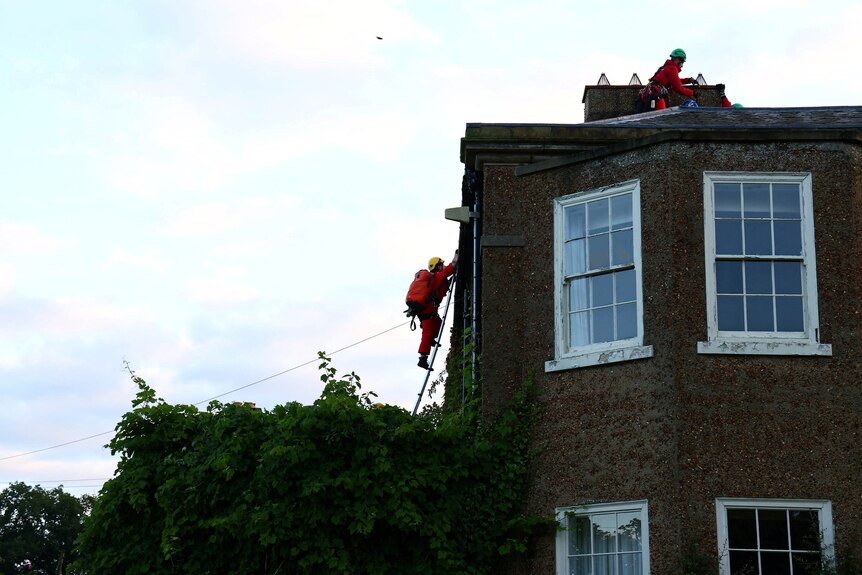 A person wearin orange climbing up a ladder on the side of a building during the day with another atop