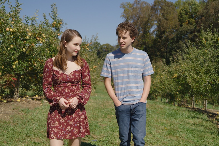 A teenage girl and a young man walk together through an orchard, standing awkwardly apart but seeming interested in each other