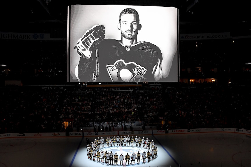 A big screen shows a black and white picture of an ice hockey player, while two teams gather on the ice below.