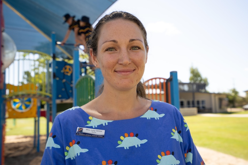Teacher smiling at the camera with kids playing on a playground in the background