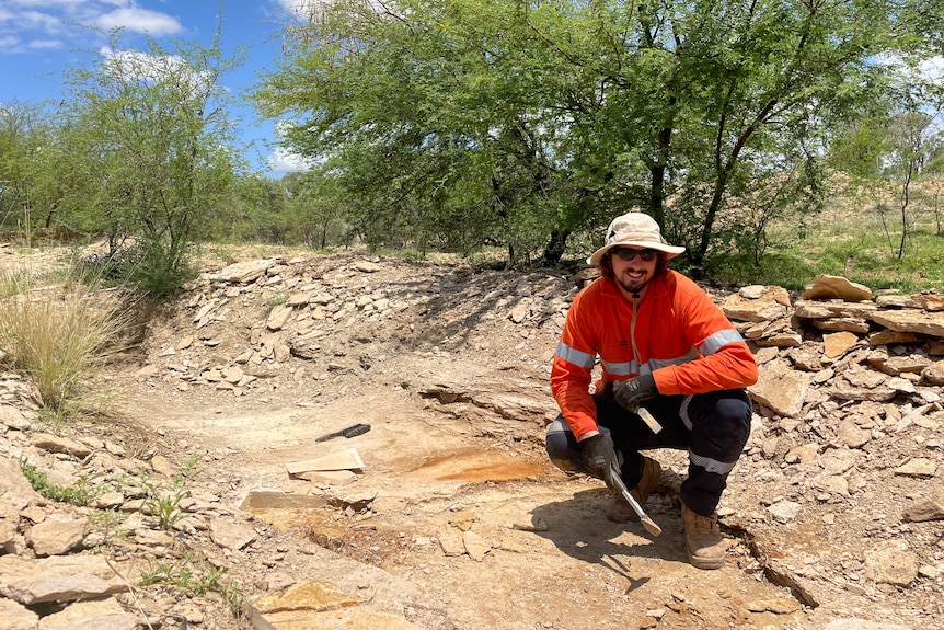 Man in hi vis shirt and hat squats in a dirt pit holding a hammer