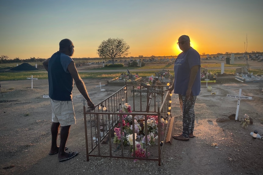 Two people stand on either side of a grave, it has a knee-high wrought iron fence and flowers.