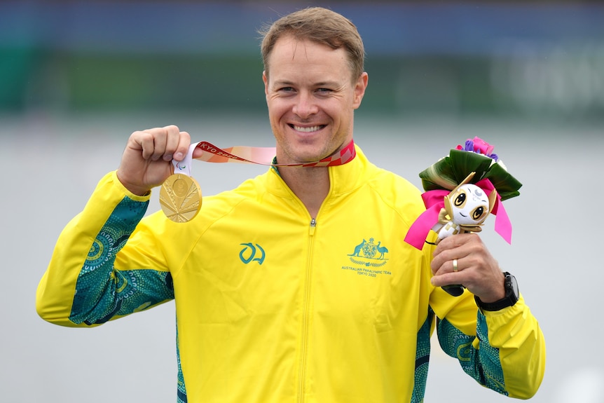 An Australian male Paralympic kayaker holds his gold medal in his right hand as she smiles.