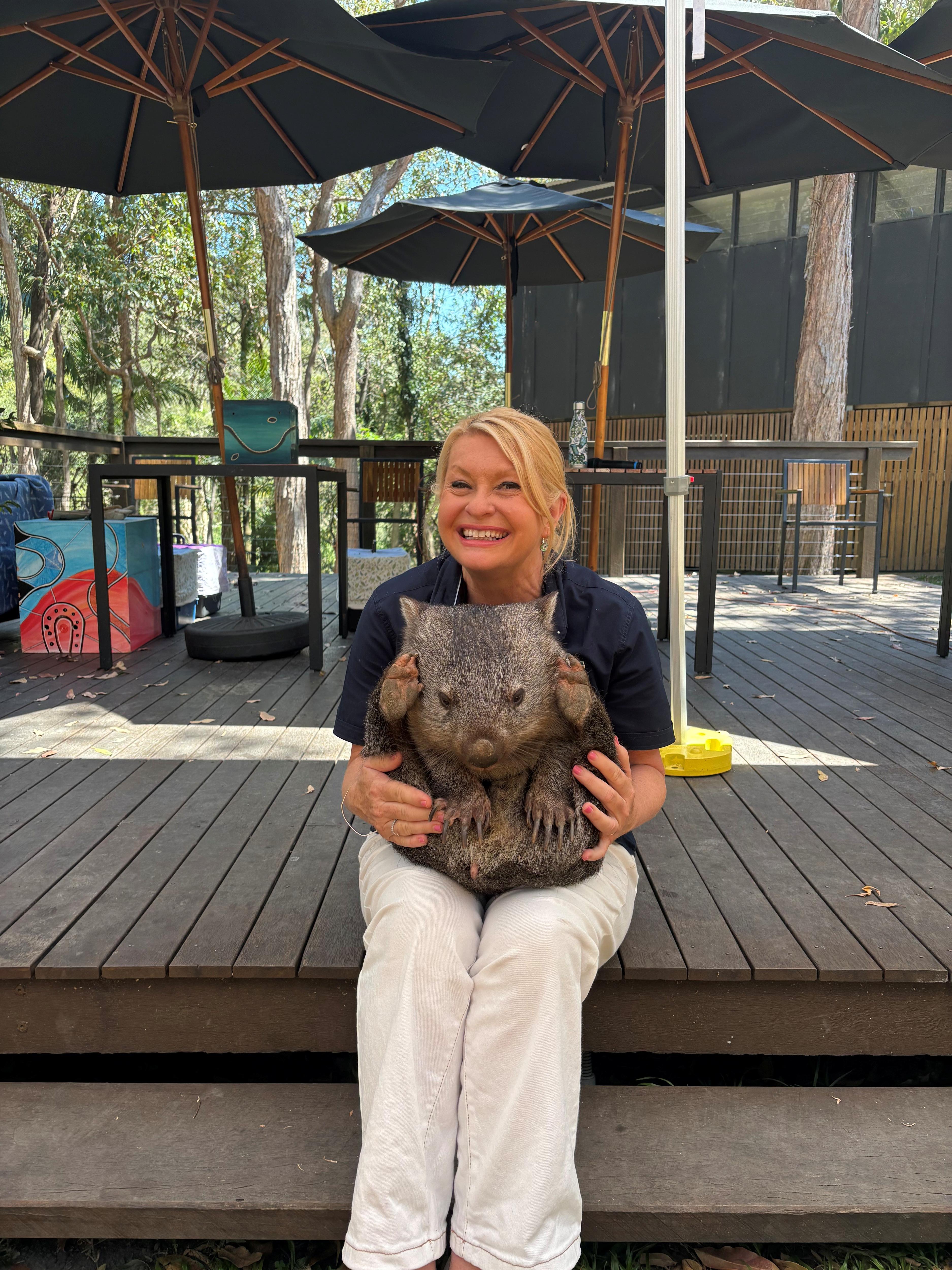A blonde woman smiles at the camera with an adult wombat in her lap.