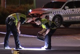 Two police officers bend examining a road cordoned off with police tape with a police car parked at the back of the shot.