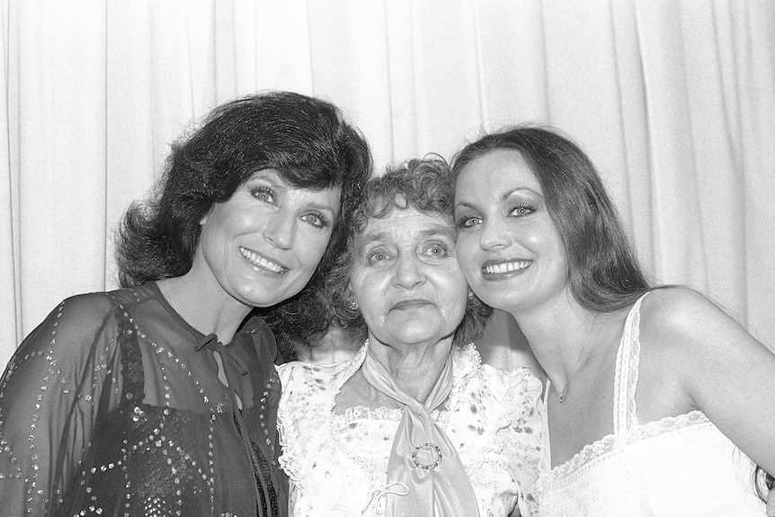 black and white image of loretta, in a see-three jewelled top, arm around her mother, with sister in white on the other side