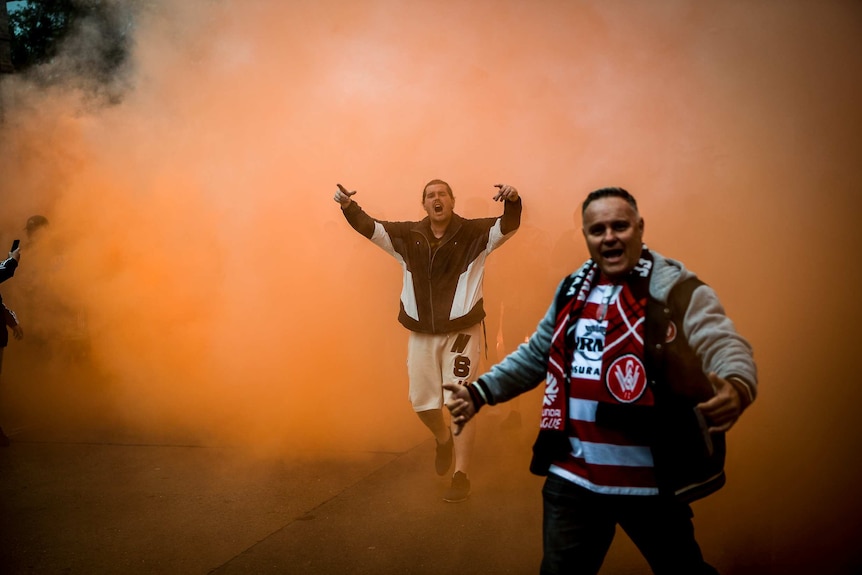 Two men walk through orange flare smoke yelling at the camera.