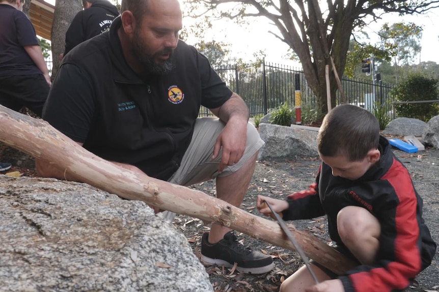 An middle-aged man crouches down next to a child who is using a tool to carve a tree branch leaning on a rock