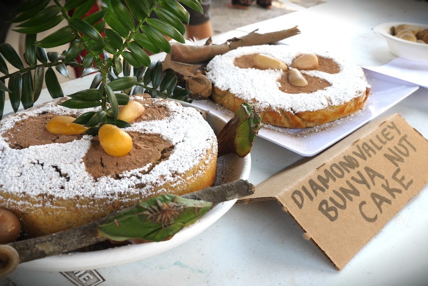 Two cakes covered in icing sugar on a table next to a small sign saying 'Diamond Valley Bunya Nut Cake' next to them.